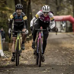 Dwie kolarki ścigają się na trasie, gdzie szosa miesza się z gravel. Uśmiechnięte zawodniczki w kaskach i strojach sportowych pędzą po leśnej ścieżce.