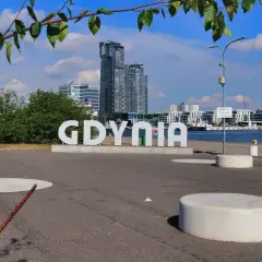 Wielkie litery "GDYNIA" na tle nowoczesnej architektury i żaglowca. Widok na port, idealny na spacer po gdynia stare miasto.