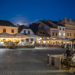 Wieczorny rynek w Kazimierzu Dolnym tętni życiem. Ludzie siedzą przy stolikach pod parasolami, delektując się posiłkami i atmosferą.