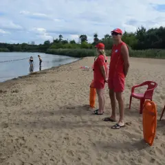 Ratownicy czuwają nad bezpieczeństwem na piaszczystym **białobrzegu**. W wodzie widać kąpiących się, a na piasku stoją dwa pomarańczowe koła ratunkowe.