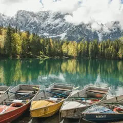 Łodzie czekają nad jeziorem w Tarvisio, odbijając się w spokojnej wodzie. W tle las i ośnieżone szczyty gór.