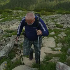 Mężczyzna w kratkach i okularach wspina się po kamienistej ścieżce, używając kijków do chodzenia.