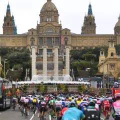 Kolarze w barwnych strojach pędzą ulicą przed monumentalnym budynkiem z fontannami, przygotowując się do etapu Tour de France.