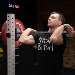 Mężczyzna przygotowuje się do przysiadu ze sztangą z przodu. Na koszulce widnieje napis "#FOMOBITCH".
