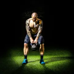 Mężczyzna z tatuażami trzyma kettlebell na tle ciemności i zielonej trawy.