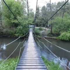 Drewniany most wiszący nad rzeką, otoczony zielenią. W oddali widać samochody, a całość tworzy malowniczy krajobraz w okolicach Celestynowa.