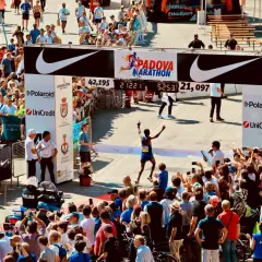Biegacz przekracza metę maratonu w Padwie, świętując zwycięstwo. Tłum kibiców i zawodników dopingują na trasie.