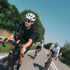 Trzech kolarzy w białych kaskach jedzie w kolumnie rowerowej po asfaltowej drodze, otoczonej zielenią.