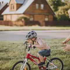 Mama uczy dziecko jeździć na rowerze. Chłopiec w kasku z radością pedałuje, a mama z uśmiechem go wspiera.