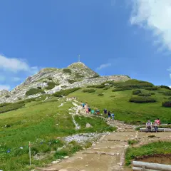 Tłum turystów wspina się kamienistym szlakiem na Giewont. Widok na szczyt z krzyżem i zielone zbocza Tatr.