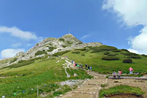 Tłum turystów wspina się kamienistym szlakiem na Giewont. Widok na szczyt z krzyżem i zielone zbocza Tatr.
