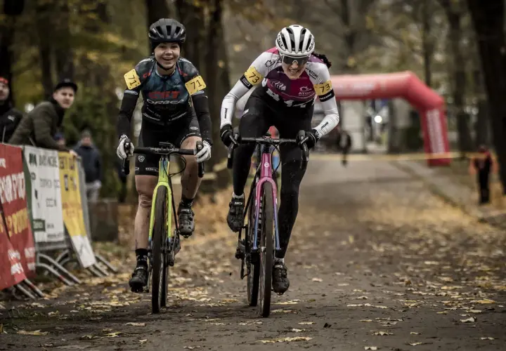 Dwie kolarki ścigają się na trasie, gdzie szosa miesza się z gravel. Uśmiechnięte zawodniczki w kaskach i strojach sportowych pędzą po leśnej ścieżce.