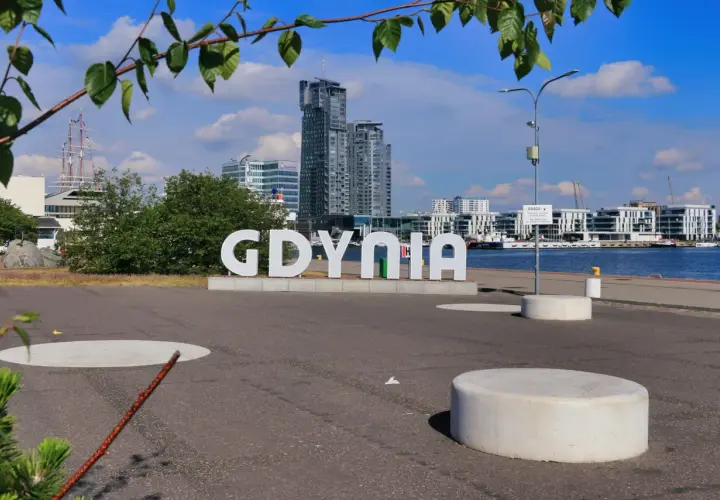 Wielkie litery "GDYNIA" na tle nowoczesnej architektury i żaglowca. Widok na port, idealny na spacer po gdynia stare miasto.