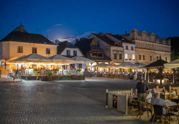 Wieczorny rynek w Kazimierzu Dolnym tętni życiem. Ludzie siedzą przy stolikach pod parasolami, delektując się posiłkami i atmosferą.