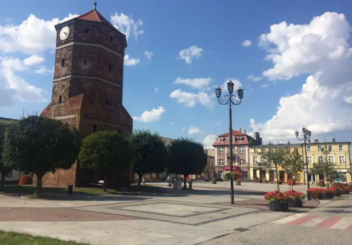 Wieża zegarowa w Barcinie góruje nad rynkiem, gdzie kwitną czerwone kwiaty.