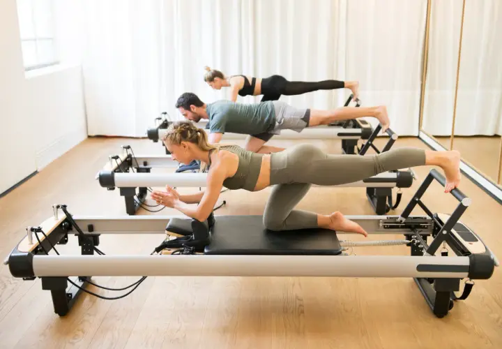 Trzy osoby ćwiczą na reformerach. To świetny sposób, by dowiedzieć się, pilates co to jest i jak poprawić sylwetkę.