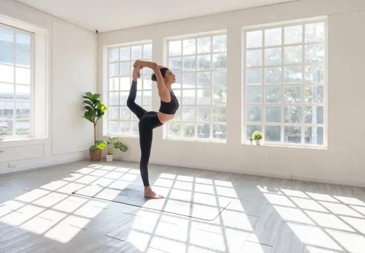 Kobieta w czarnym stroju wykonuje pozycję Natarajasana podczas sesji vinyasa jogi. Światło wpada przez okna.