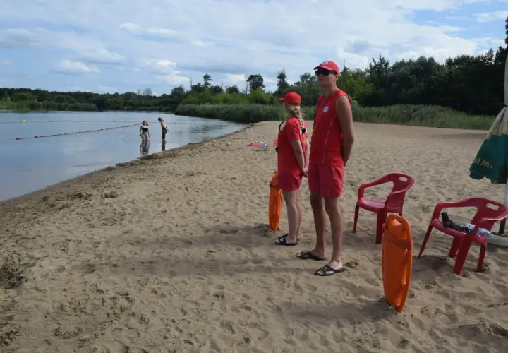 Ratownicy czuwają nad bezpieczeństwem na piaszczystym **białobrzegu**. W wodzie widać kąpiących się, a na piasku stoją dwa pomarańczowe koła ratunkowe.