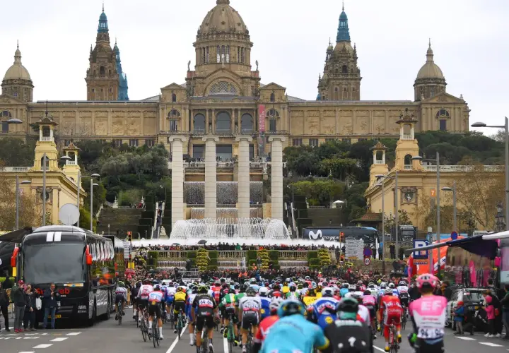 Kolarze w barwnych strojach pędzą ulicą przed monumentalnym budynkiem z fontannami, przygotowując się do etapu Tour de France.