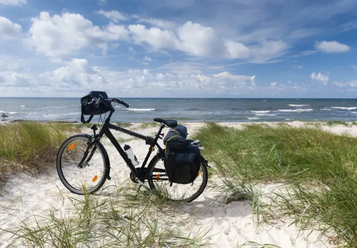 Rower z sakwami na piaszczystej plaży, gotowy na odkrywanie tras rowerowych nad morzem. Błękitne niebo i fale tworzą idealne tło.