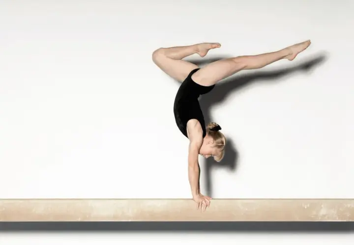 Gimnastka w czarnym stroju wykonuje precyzyjny handstand na równoważni, z nogami w powietrzu.