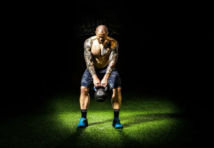 Mężczyzna z tatuażami trzyma kettlebell na tle ciemności i zielonej trawy.