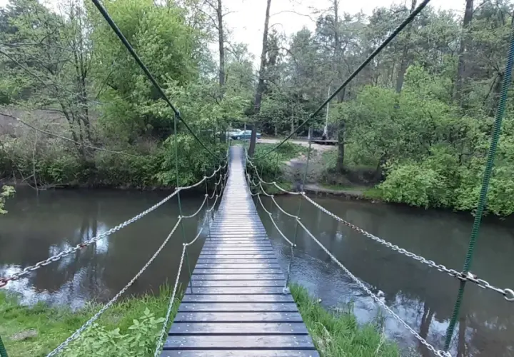 Drewniany most wiszący nad rzeką, otoczony zielenią. W oddali widać samochody, a całość tworzy malowniczy krajobraz w okolicach Celestynowa.