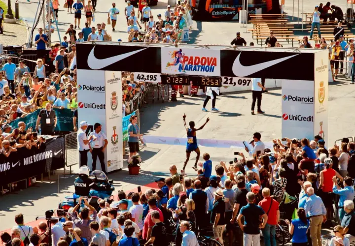 Biegacz przekracza metę maratonu w Padwie, świętując zwycięstwo. Tłum kibiców i zawodników dopingują na trasie.