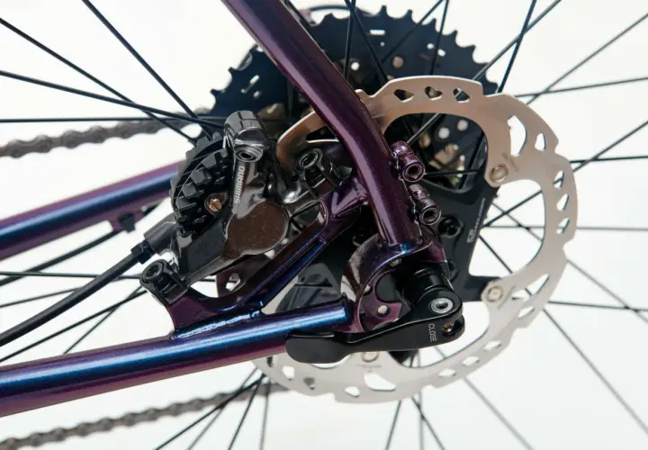 Błyszcząca, fioletowa rama roweru z widoczną sztywną osią, przerzutką Shimano i tarczą hamulcową.
