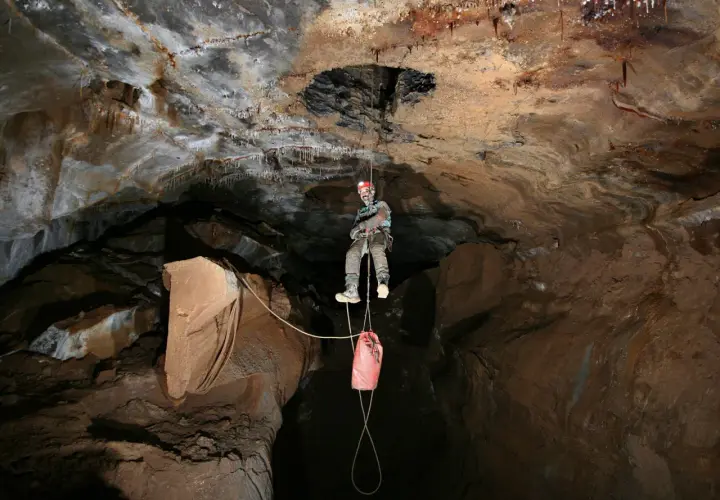 Odważny speleolog wspinający się po linie w jaskini, z workiem na sprzęt zwisającym poniżej.