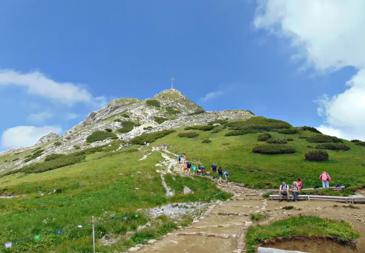 Tłum turystów wspina się kamienistym szlakiem na Giewont. Widok na szczyt z krzyżem i zielone zbocza Tatr.
