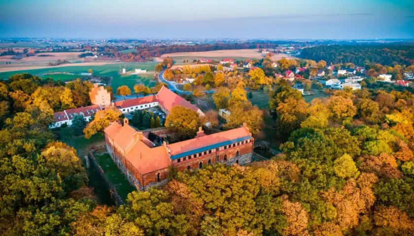 Zamek Bierzgłowski otoczony jesiennym lasem, z czerwonymi dachami i ceglanymi murami, w blasku zachodzącego słońca.