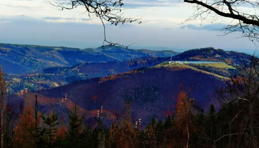Widok na górzyste pasmo z drzewami w jesiennych barwach. Na szczycie jednego ze wzgórz widać budynki i zbiornik wodny. W oddali majaczy maszt telekomunikacyjny, przypominający czupel.