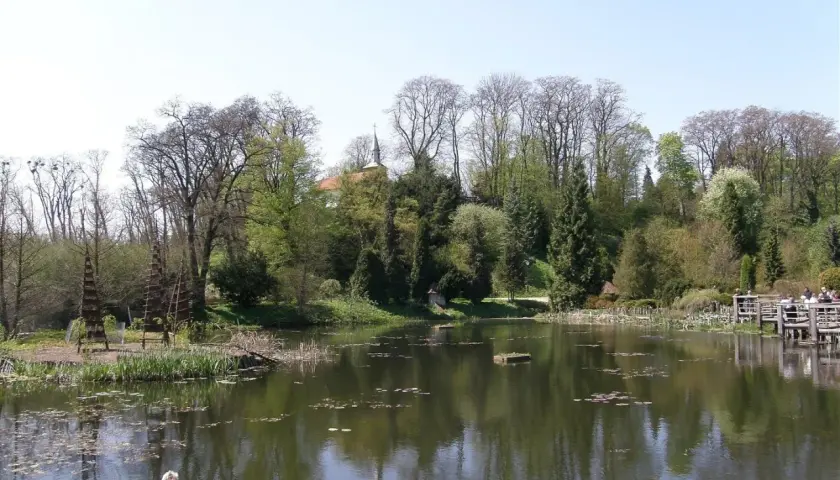 Malowniczy staw w Bolestraszycach, otoczony drzewami i zielenią. Widoczny fragment pałacu i drewniany pomost, idealne miejsce na relaks i podziwianie atrakcji.