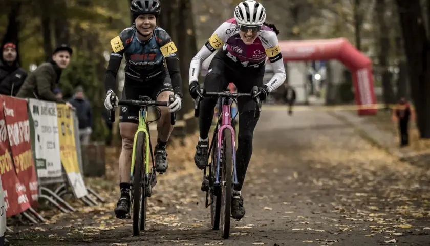 Dwie kolarki ścigają się na trasie, gdzie szosa miesza się z gravel. Uśmiechnięte zawodniczki w kaskach i strojach sportowych pędzą po leśnej ścieżce.