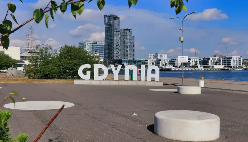 Wielkie litery "GDYNIA" na tle nowoczesnej architektury i żaglowca. Widok na port, idealny na spacer po gdynia stare miasto.