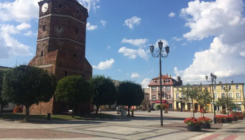 Wieża zegarowa w Barcinie góruje nad rynkiem, gdzie kwitną czerwone kwiaty.