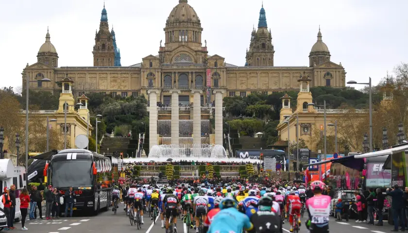 Kolarze w barwnych strojach pędzą ulicą przed monumentalnym budynkiem z fontannami, przygotowując się do etapu Tour de France.