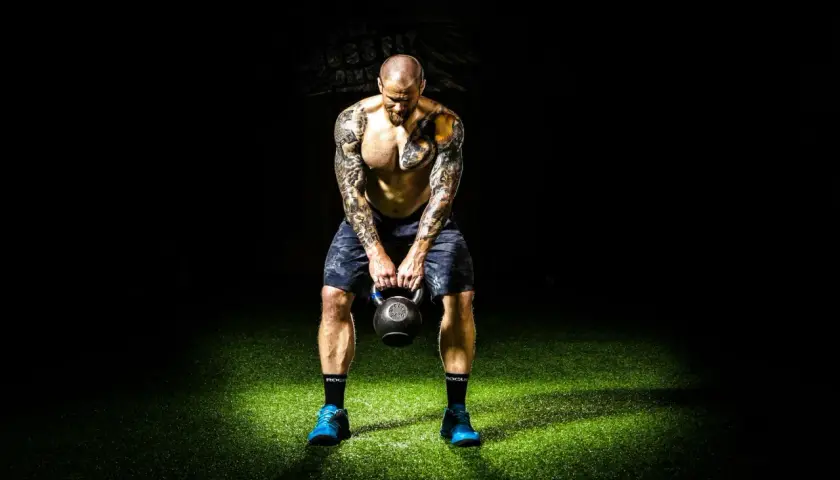 Mężczyzna z tatuażami trzyma kettlebell na tle ciemności i zielonej trawy.