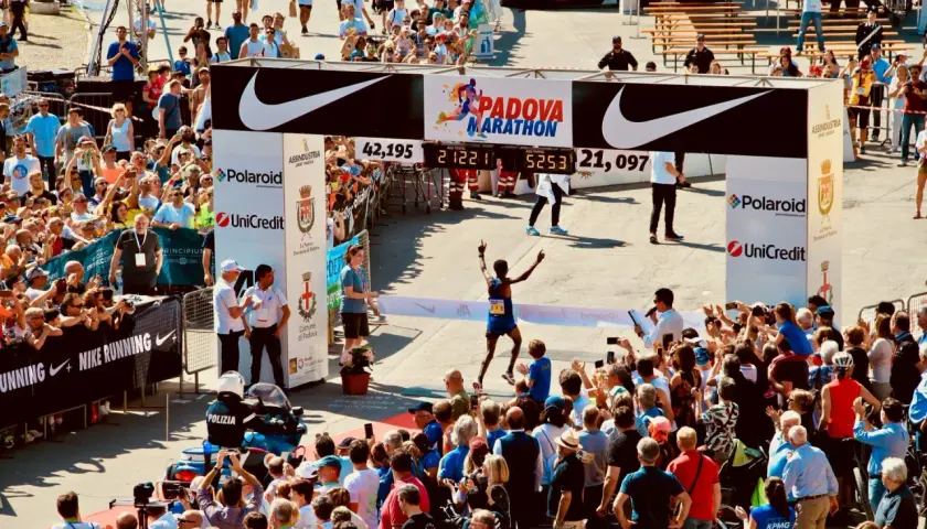 Biegacz przekracza metę maratonu w Padwie, świętując zwycięstwo. Tłum kibiców i zawodników dopingują na trasie.