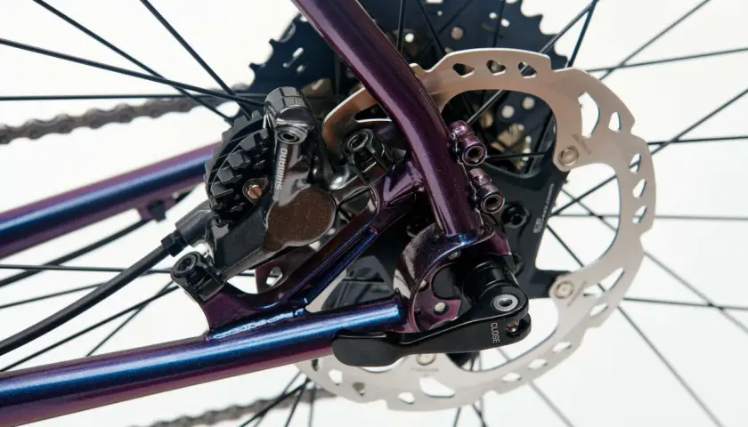 Błyszcząca, fioletowa rama roweru z widoczną sztywną osią, przerzutką Shimano i tarczą hamulcową.