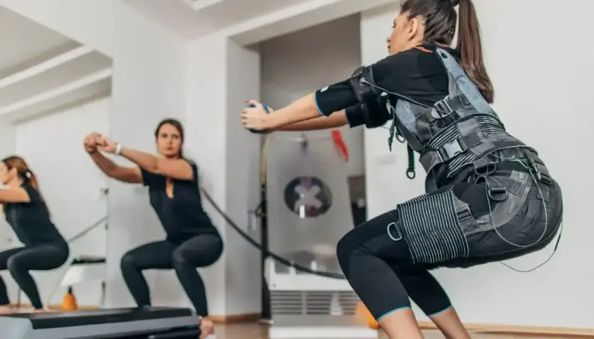 Kobiety w kamizelkach EMS wykonują przysiady. Trening z elektrostymulacją mięśni (EMS) to przyszłość fitnessu.