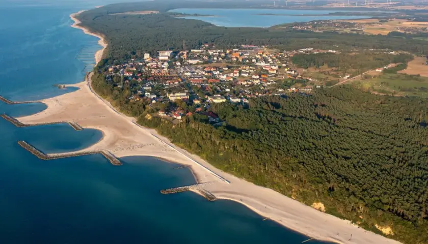 Widok z lotu ptaka na Jarosławiec, nadmorską miejscowość z piaszczystą plażą, lasami i zabudowaniami.