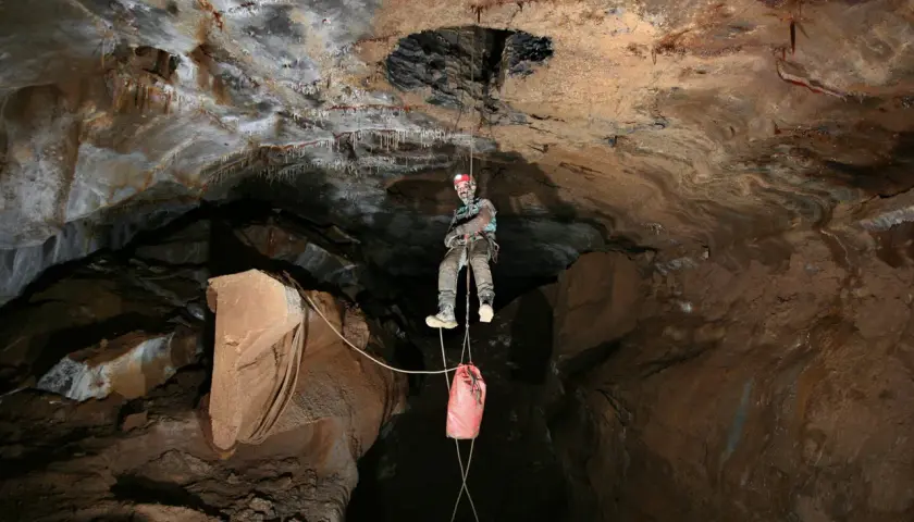 Odważny speleolog wspinający się po linie w jaskini, z workiem na sprzęt zwisającym poniżej.