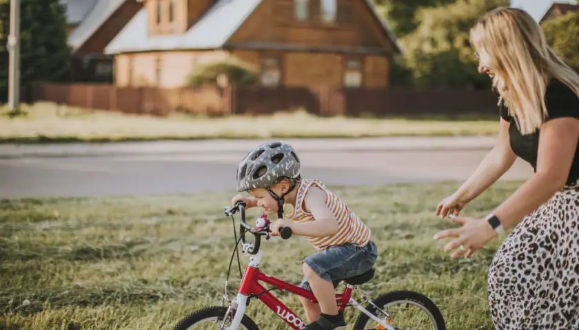 Mama uczy dziecko jeździć na rowerze. Chłopiec w kasku z radością pedałuje, a mama z uśmiechem go wspiera.