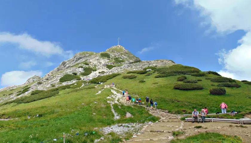 Tłum turystów wspina się kamienistym szlakiem na Giewont. Widok na szczyt z krzyżem i zielone zbocza Tatr.