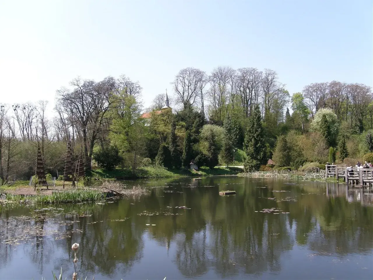 Malowniczy staw w Bolestraszycach, otoczony drzewami i zielenią. Widoczny fragment pałacu i drewniany pomost, idealne miejsce na relaks i podziwianie atrakcji.