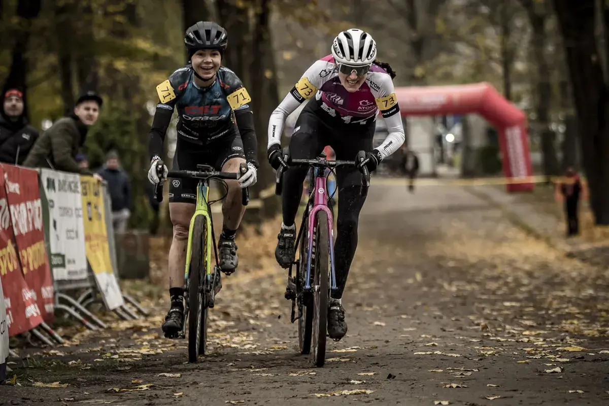 Dwie kolarki ścigają się na trasie, gdzie szosa miesza się z gravel. Uśmiechnięte zawodniczki w kaskach i strojach sportowych pędzą po leśnej ścieżce.