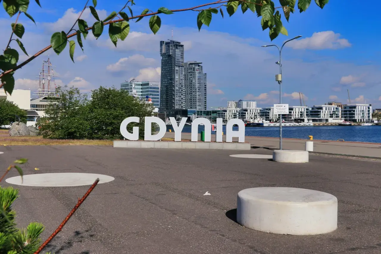 Wielkie litery "GDYNIA" na tle nowoczesnej architektury i żaglowca. Widok na port, idealny na spacer po gdynia stare miasto.