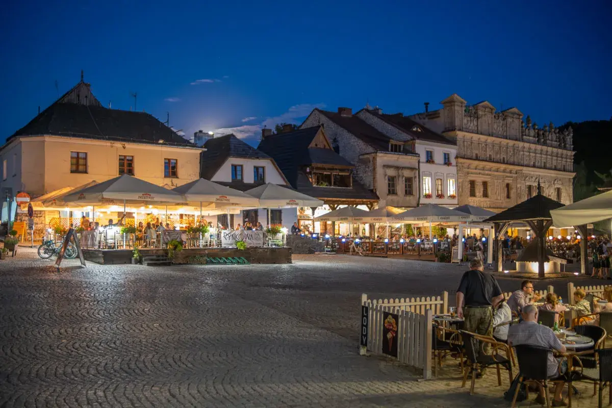 Wieczorny rynek w Kazimierzu Dolnym tętni życiem. Ludzie siedzą przy stolikach pod parasolami, delektując się posiłkami i atmosferą.